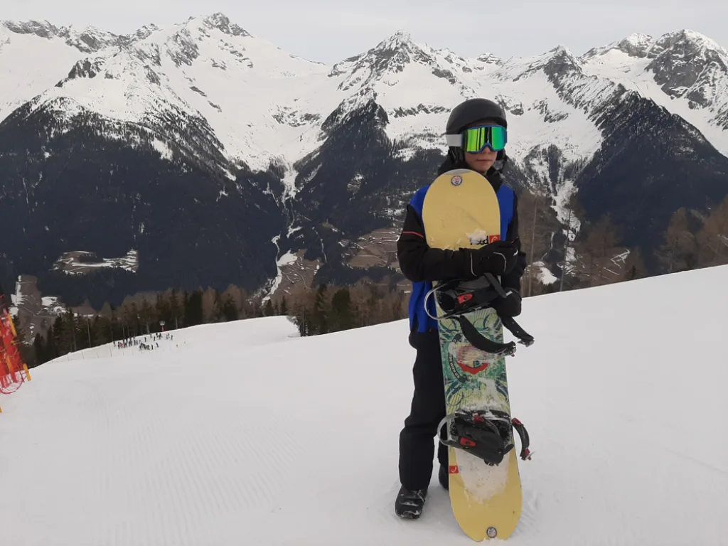 Ein Jugendlicher hält sein Snowboard hoch, dahinter die verschneiten Berge.